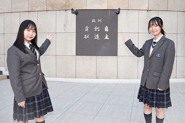 『自主 創造 貢献』の校訓の前で微笑む、KさんとAさんの写真です